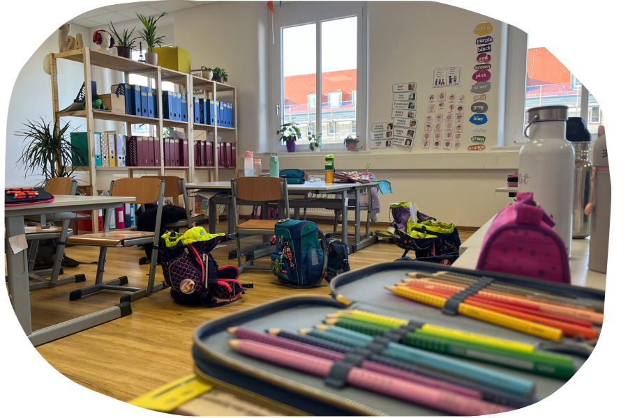 Heller Klassenraum mit Tischen, Stühlen, Schulranzen und farbigen Stiften auf einem Schreibtisch.